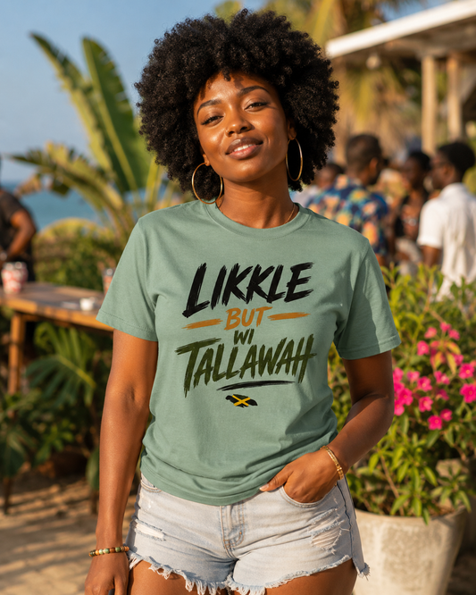 A woman with an afro hairstyle wearing a sage green graphic t-shirt that reads 'LIKKLE BUT IN TALLAWAH' in black and yellow text, paired with light denim shorts, standing outdoors in a sunny, tropical environment.