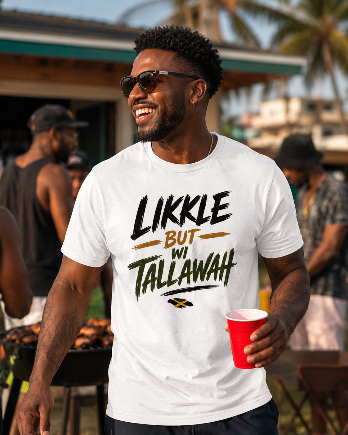 A man wearing a white graphic t-shirt with the text 'LIKKE BUT WI TALLAWAH' in black and green, holding a red cup at an outdoor barbecue.